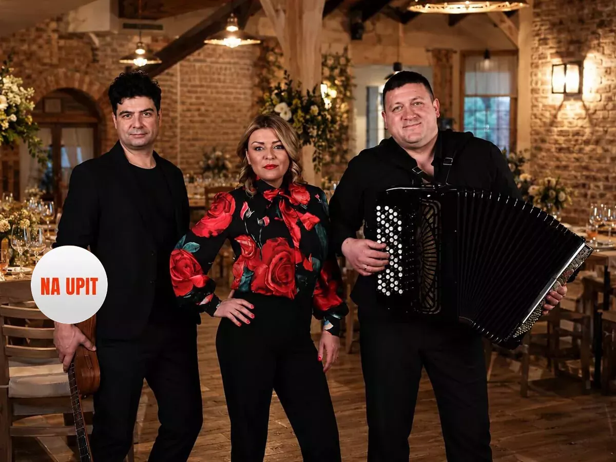 Članovi benda A&A Bend poziraju ispred sive zidne pozadine — muškarac sa gitarom, žena u bluzi sa crvenim ružama i muškarac sa harmonikom. Tekst na slici: "A&A Bend", "Već od 700€