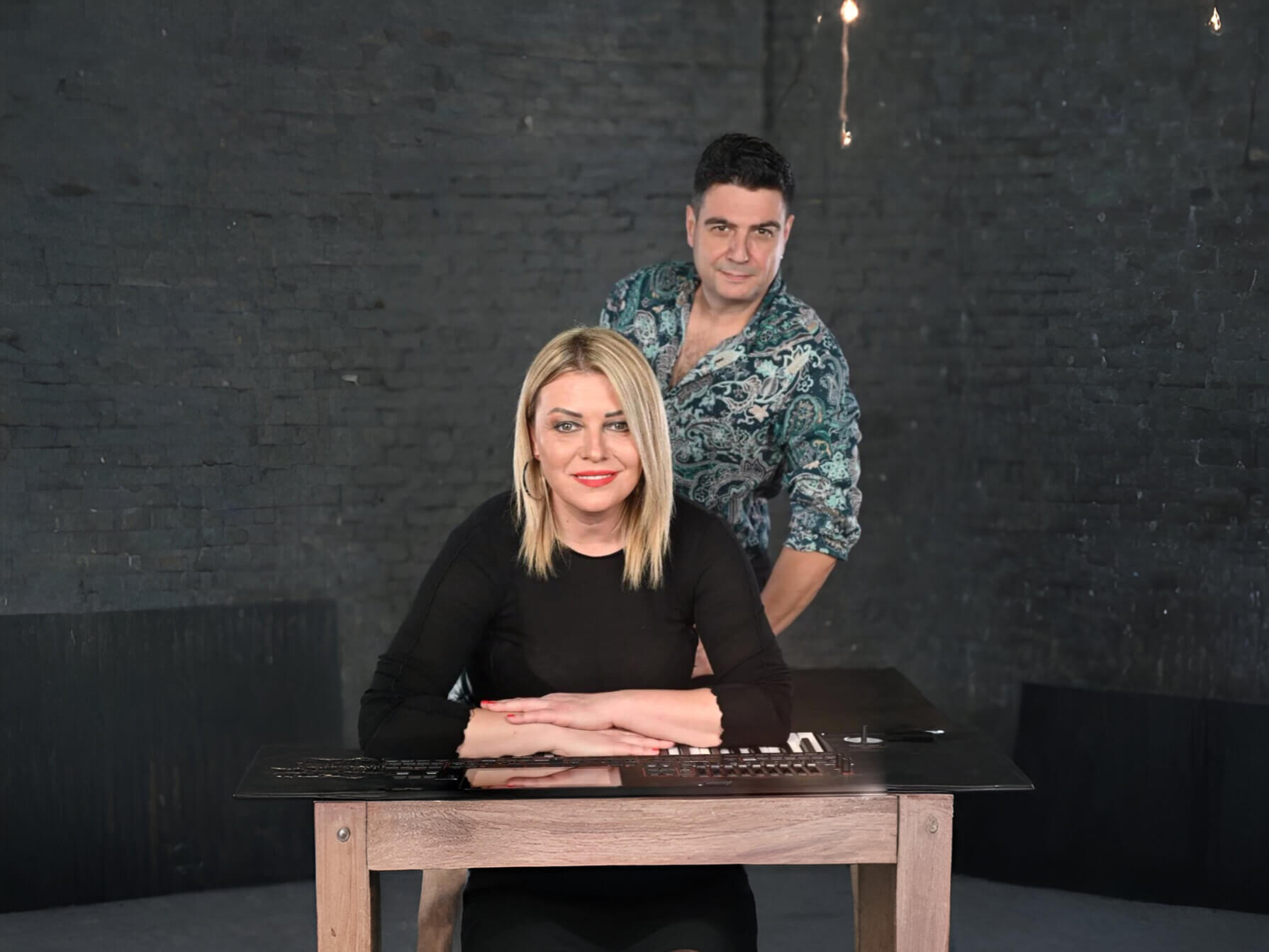 žena sedi za klavijaturoma u crnoj majici muškarac stoji iza nje u šarenoj košulji