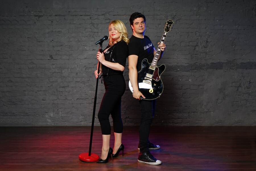 muškarac drži gitaru a žena mikrofon, oboje u crnom, leđima okrenuti jedno drugom