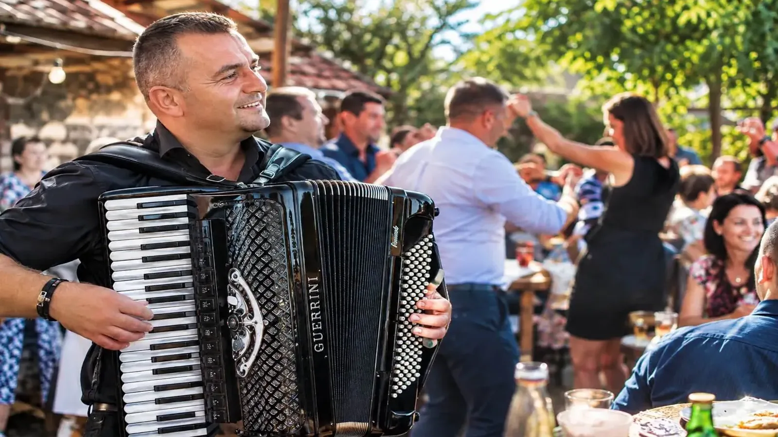 Harmonikaš svira harmoniku na veselju na otvorenom u Srbiji, dok gosti plešu i druže se za stolovima.