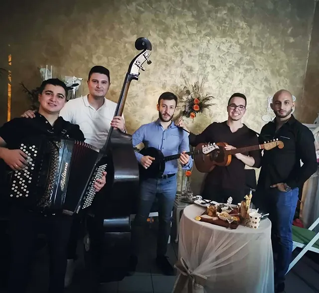 Tamburaški bend za proslave pozira u restoranu, muzičari sa harmonikom, kontrabasom i tamburama ispred dekorisanog stola sa hranom i pićem