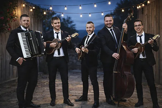Tamburaši za proslave stoje napolju u večernjem ambijentu pod lampicama, pet muzičara u crnim odelima svira tamburice, harmoniku i kontrabas, nasmejani i spremni za nastup na slavlju.